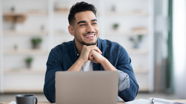 Man smiles whilst sat down at a desk with laptop open in front of him